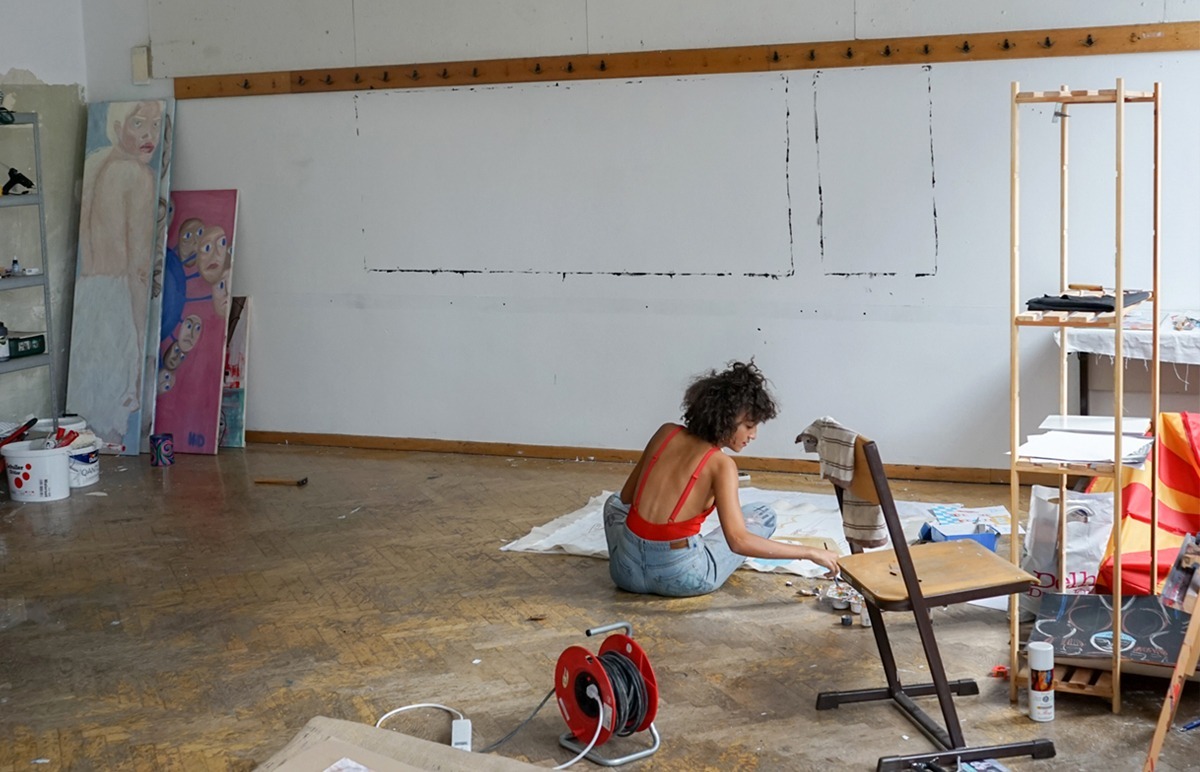 Eine Person sitzt im Atelier des Never At Home Projekts auf dem Boden und malt. Gemälde lehnen an der Wand und Farben stehen bereit.