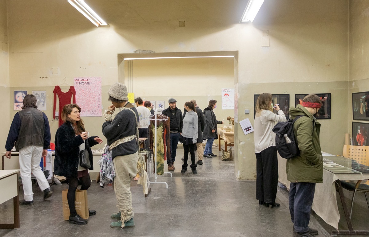 Personen laufen durch einen ehemaligen Werkraum einer alten Schule. Der Raum ist nun Schauplatz einer Ausstellung. Die Wände sind mit Bildern versehen, Tische stehen an den Wänden des Raumes, eine Kleiderstange stehe im Durchgang zwischen den Räumen.