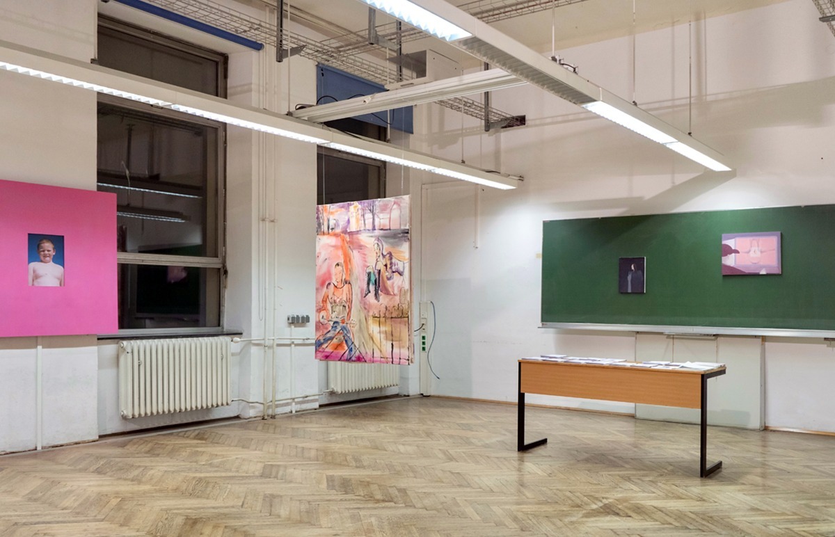 Ein ehemaliger Klassenraum ist zu einer künstlerischen Ausstellung umfunktioniert worden. Auf der rechten Seite des Bildes erinnert die Tafel und das davorstehende Lehrerpult noch an die vorherige Nutzung. Große in Rosé Töne getauchte Bilder hängen von der Decke und an der linken Seite des Bildes direkt neben der Fensterfront.