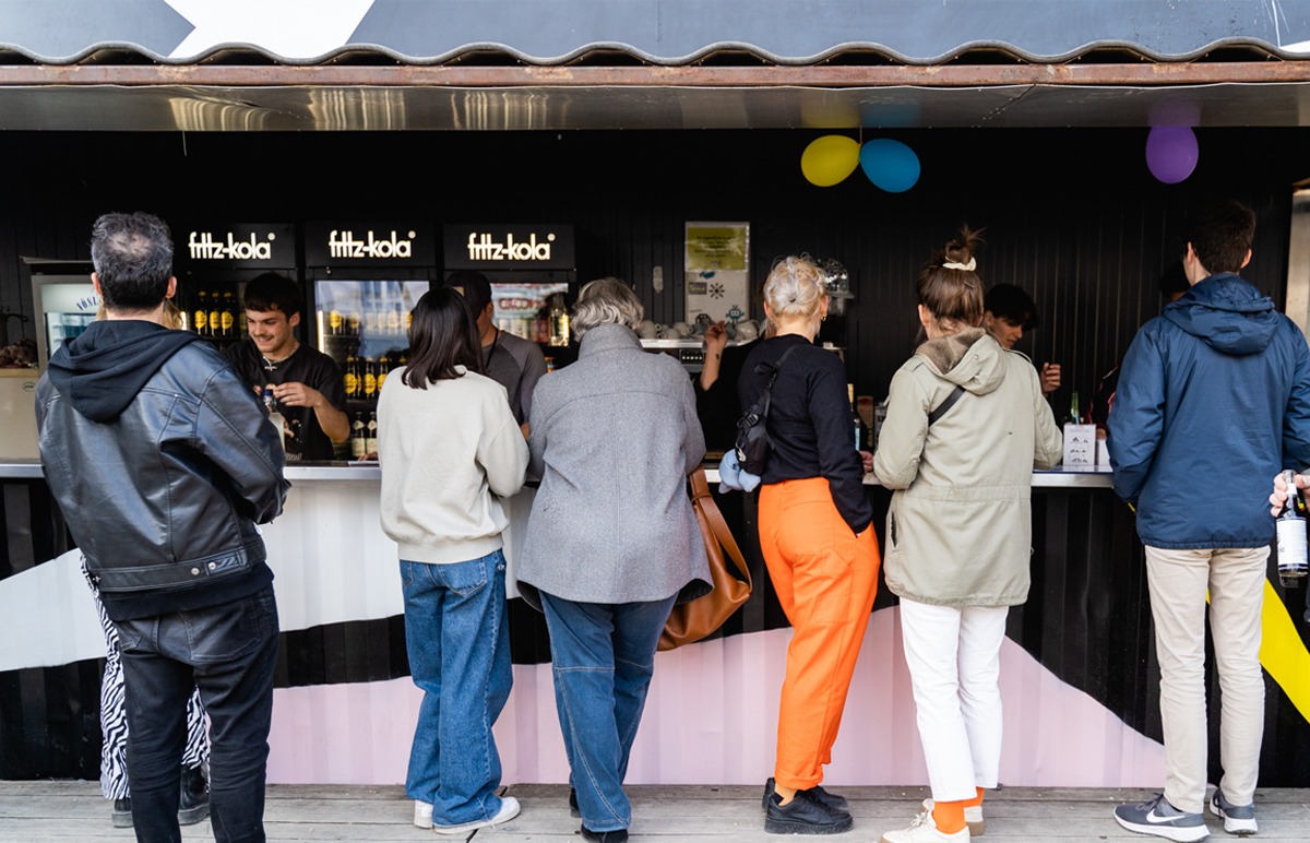 Eine bunte Bar ist zu sehen. Luftballons hängen von der Decke. Vor der Bar stehen mehre Personen um ein Bestellung aufzugeben. Sie sind dem Betrachter mit dem Rücken zugewandt. Hinter der Barr stehen mehrere Männer, die mit einem Lächeln im Gesicht die Kunden bedienen.