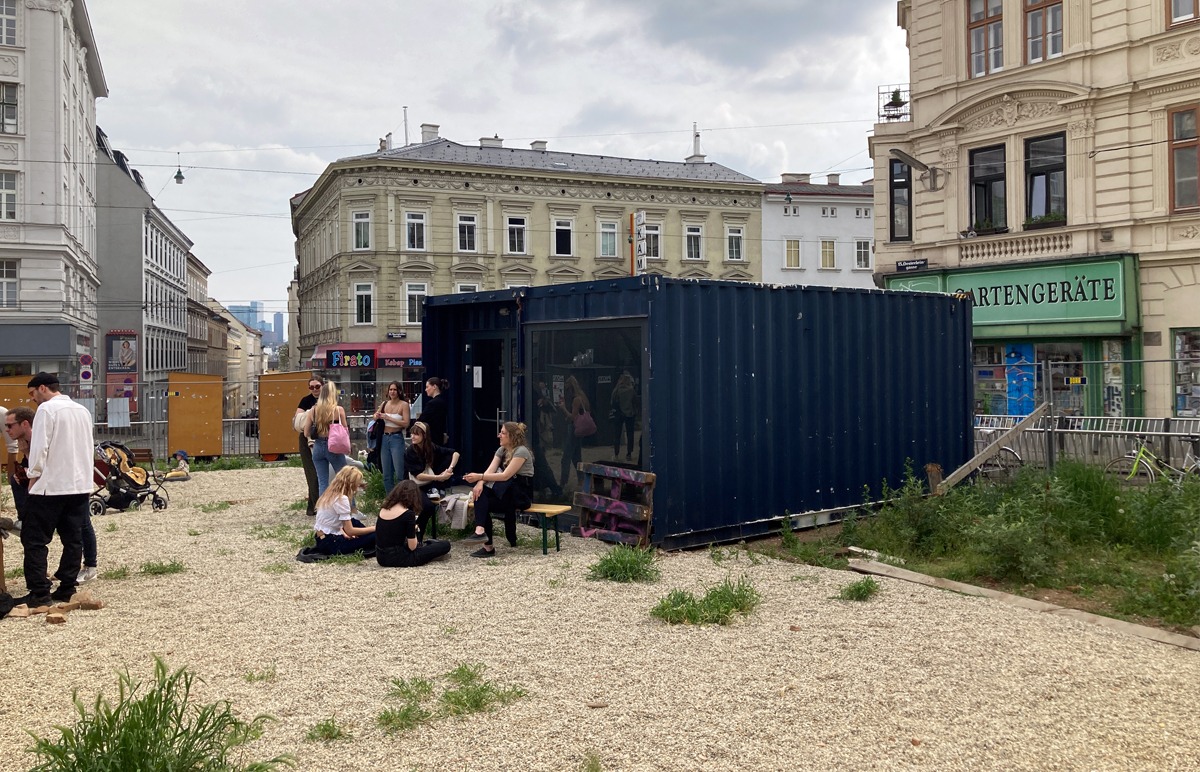 Durch den Kiesboden drücken sich kleine Plfanzen durch. Auf der Fläche steht ein dunkel blauer Container. Vor diesem sitzen mehrere Personen auf einer Bierbank.