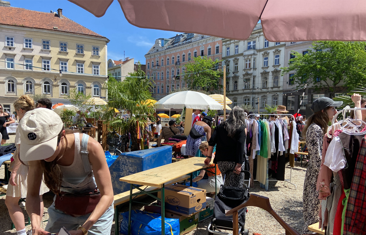 Ein Kleiderflohmarkt mit einem bunten Sortiment and Kleidungsstücken in Sonnenstrahlen gebadet mit vielen Besuchern.