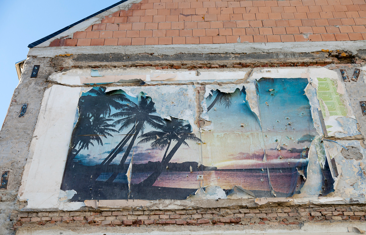 Man sieht die Überreste einer Abgerissenen Fassade. An der Wand des angrenzenden Hauses sind die Reste einer Palmentapete zu sehen.