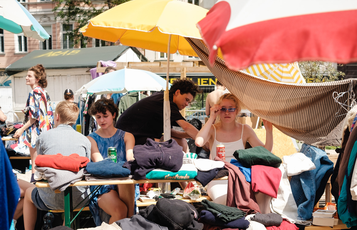 Menschen verkaufen Kleidung im Freien. Dahinter Sonnenschirme. 