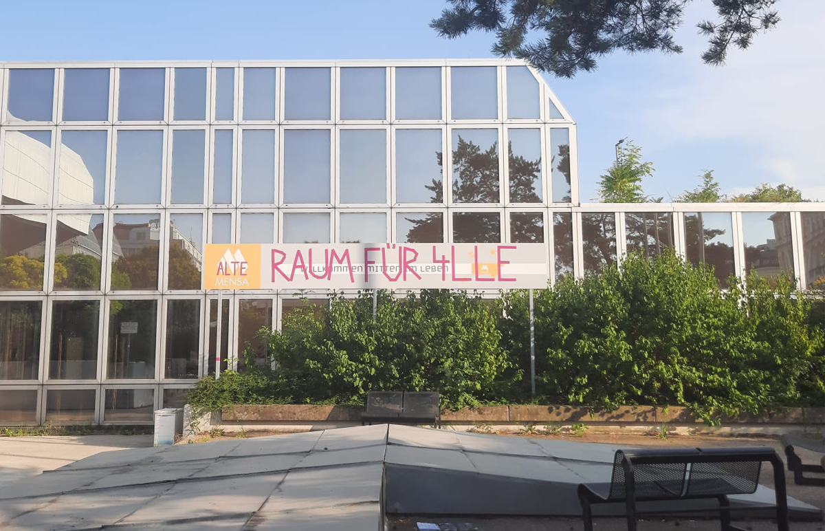 Ein Gebäude mit großer Fensterfront und einem Banner auf dem Raum für Alle geschrieben steht.