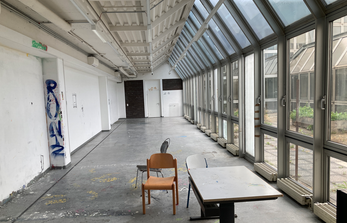 Raum mit drei Stühlen und einem Tisch, links ist eine weiße Wand mit Graffiti, rechts eine Glaswand zum Innenhof.