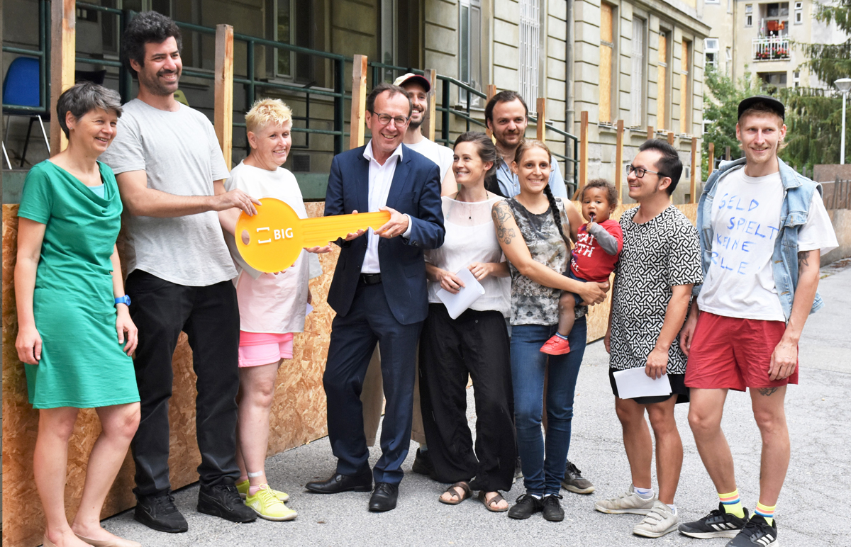 Bezirksvorsteherin Silvia Nossek Uli Fries von Kreative Räume Wien, Barbara Klampfl vom Verein Semmelweis, Wolfgang Gleissner von der BIG und weitere Personen vom Verein Semmelweis stehen vor dem Gebäude und halten einen gelben Schlüssel mit der Aufschrift BIG