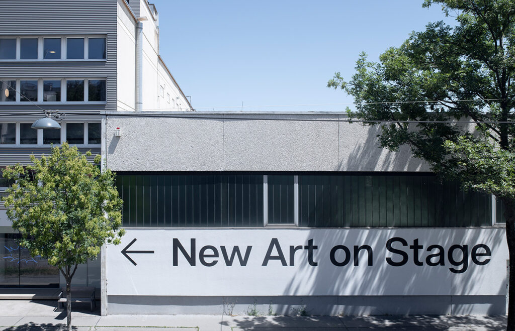 new art on stage steht mit großen Lettern mit einem schwarzen pfeil auf einer häuserfront von einem grauen bürotrakt.