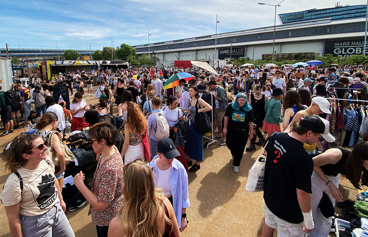 Viele Menschen im Sonnenschein auf einem Flohmarkt, im Hintergrund Gebäude.