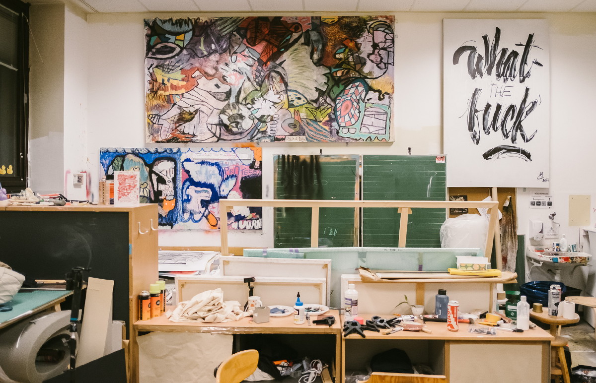 Frontaler Blick in ein Atelier des Creative Cluster, mit Arbeitsplätzen, einer Tafel (ehemaliges Klassenzimmer) und Malerei an den Wänden.