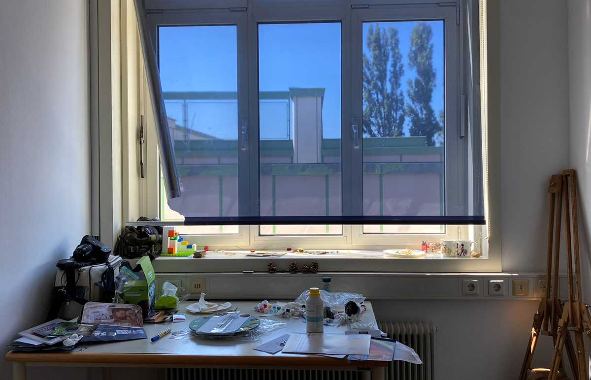 frontaler Blick in das kleine Atelier und den Arbeitsplatz einer Künstlerin, mit einer Folienjalousie, die das Fenster verdunkelt