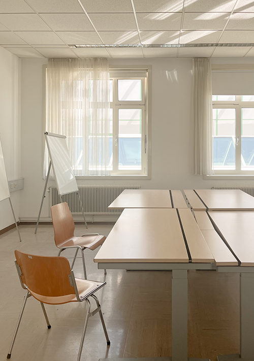 Blick Richtung Fenster in einen Raum mit vier Tischen, zwei Stühlen und Flipcharts