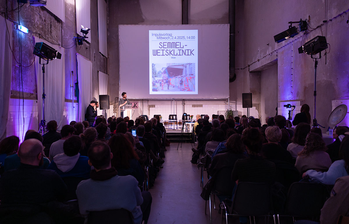 Präsentation und ein voll besetzter Raum mit Zuschauer:innnen.