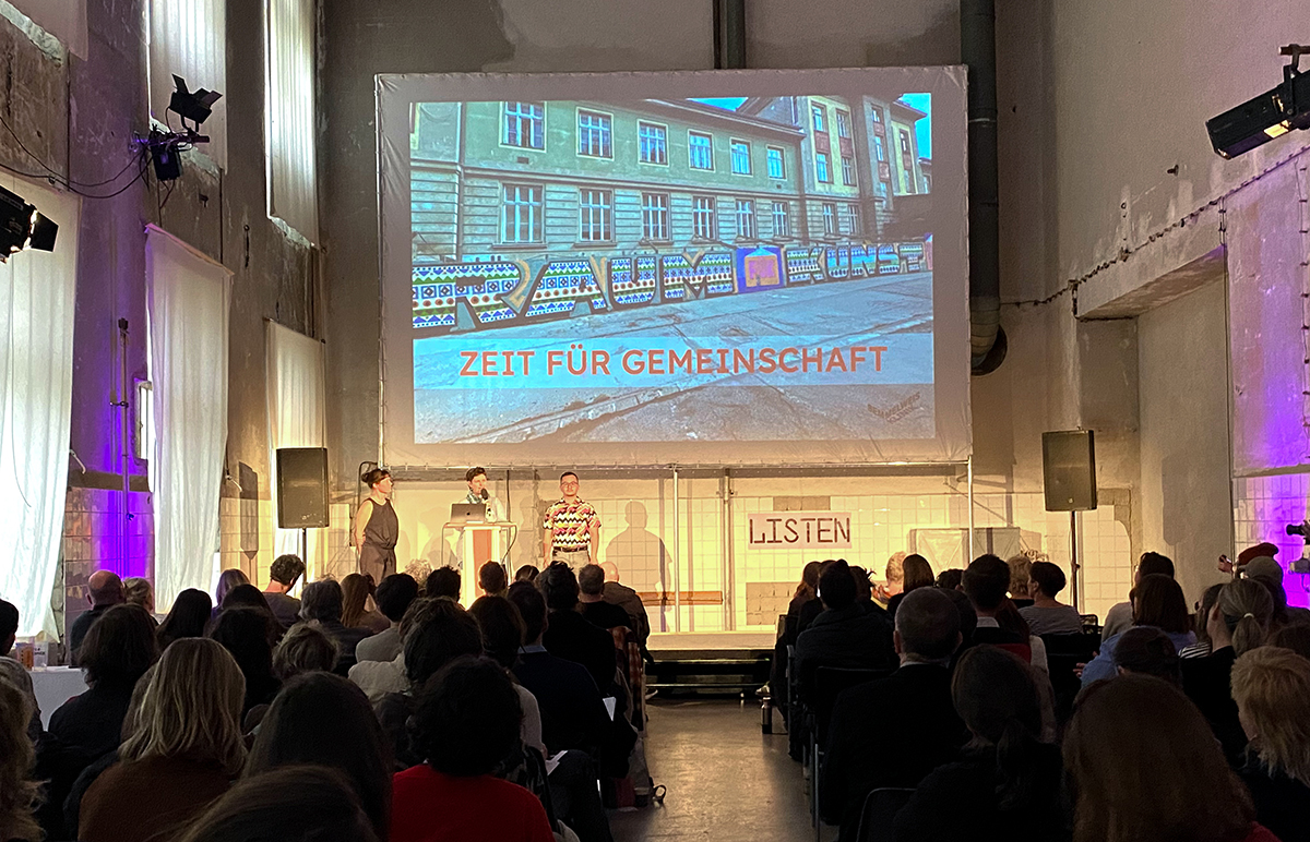 Präsentation und ein voll besetzter Raum mit Zuschauer:innnen.